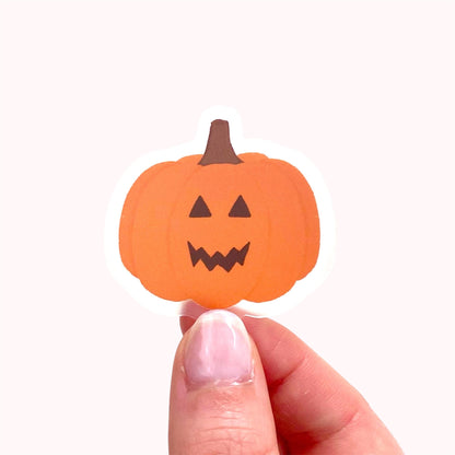 Orange jack-o-lantern pumpkin sticker held by hand against a pink background.