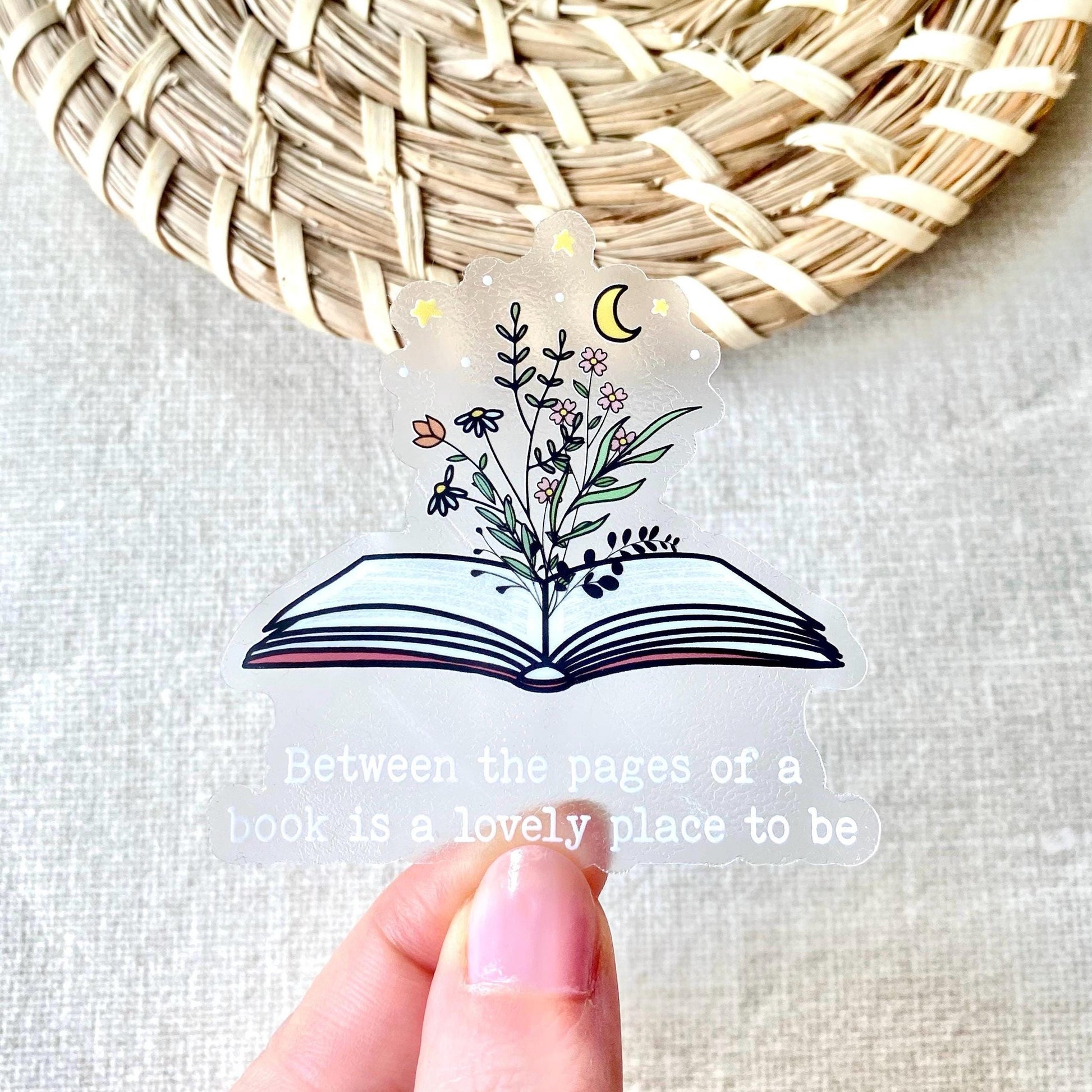 Hand holding a clear book sticker with floral design and quote, woven mat in background