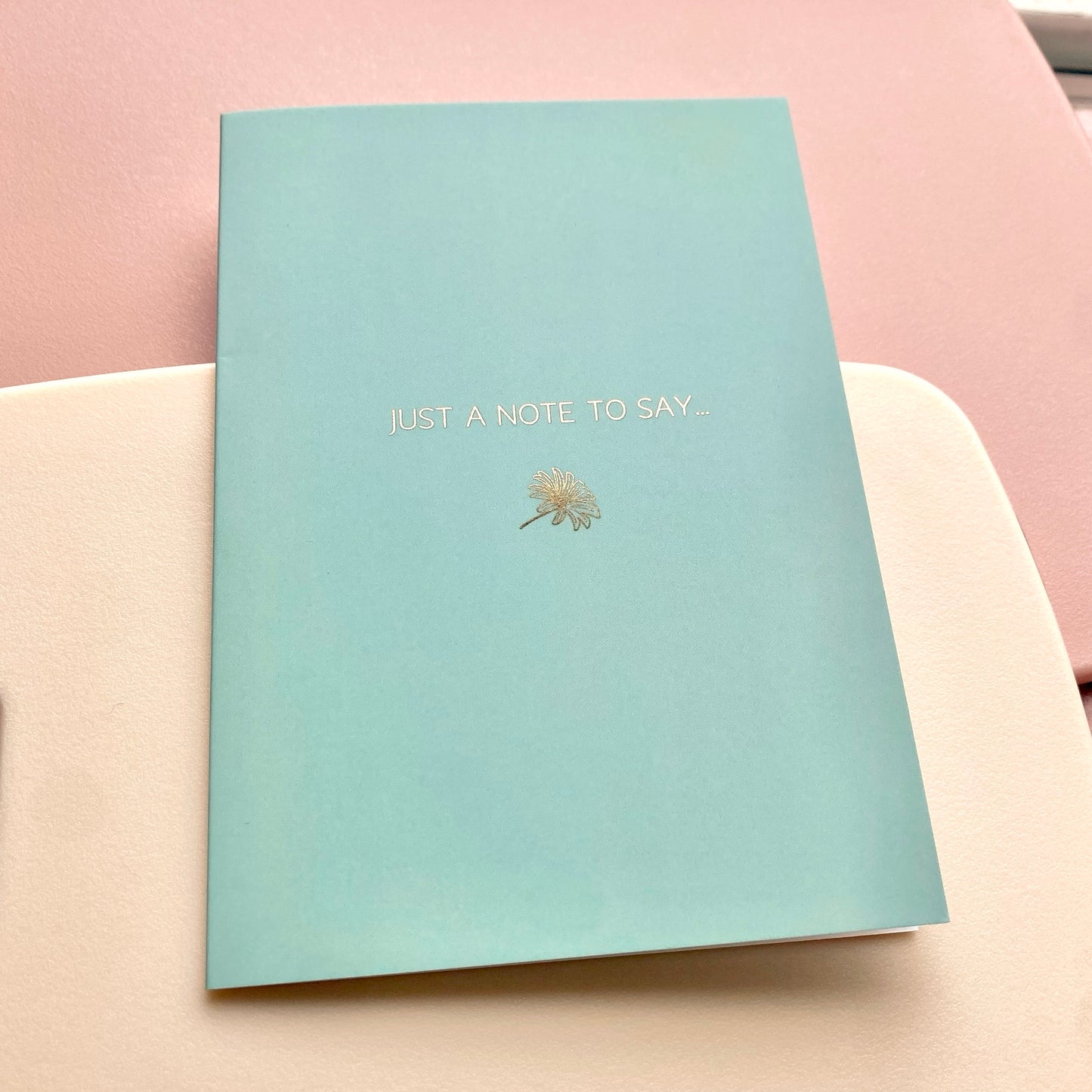 Mint blue card with gold text and daisy design, lying on a pink and cream background.