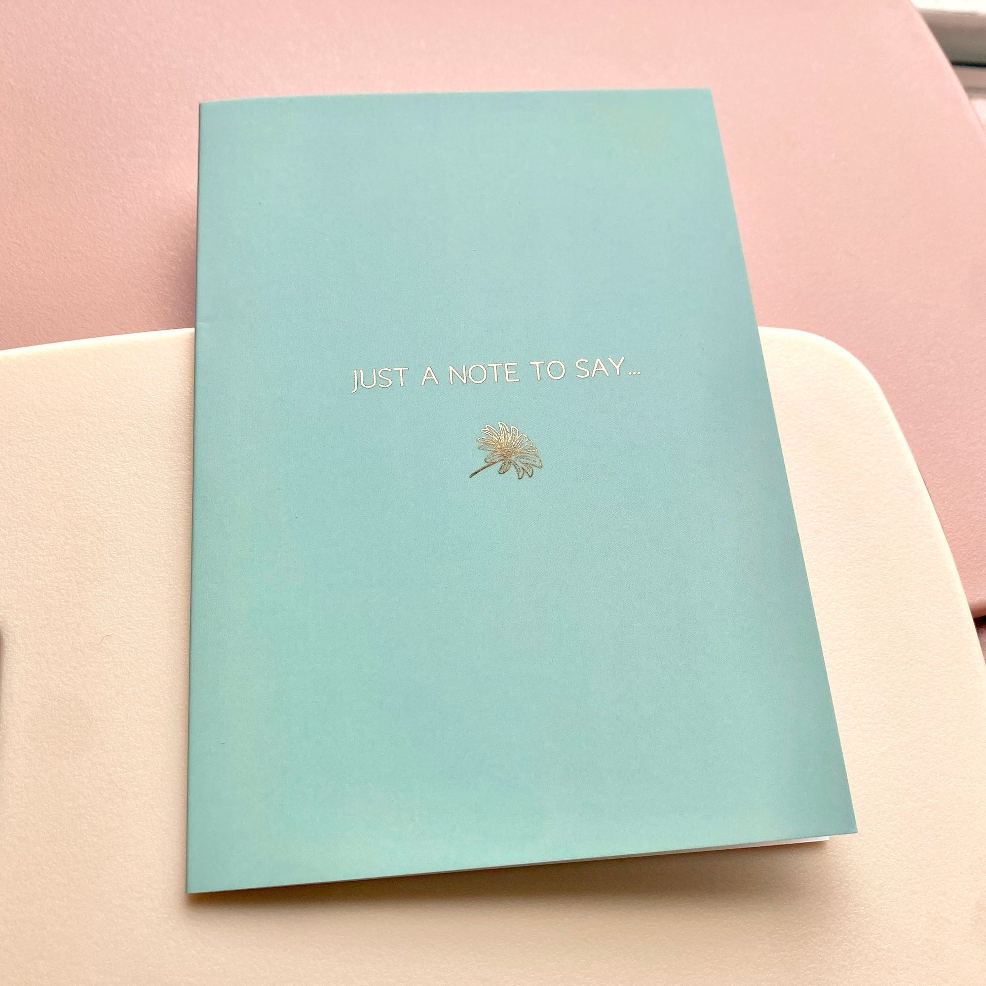 Mint blue card with gold text and daisy design, lying on a pink and cream background.