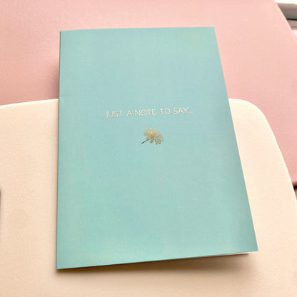 Mint blue card with gold text and daisy design, lying on a pink and cream background.