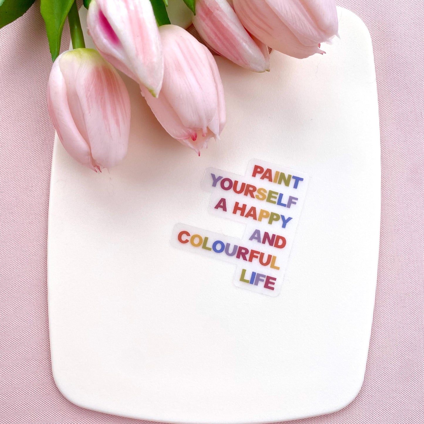 A clear sticker with motivational quote 'Paint yourself a happy and colourful life' on a white background with pink tulips