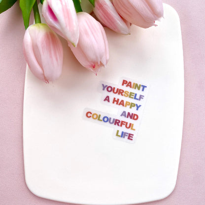 A clear sticker with motivational quote 'Paint yourself a happy and colourful life' on a white background with pink tulips