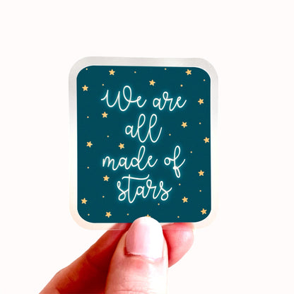 Sticker with 'We are all made of stars' text held by a hand on a white background