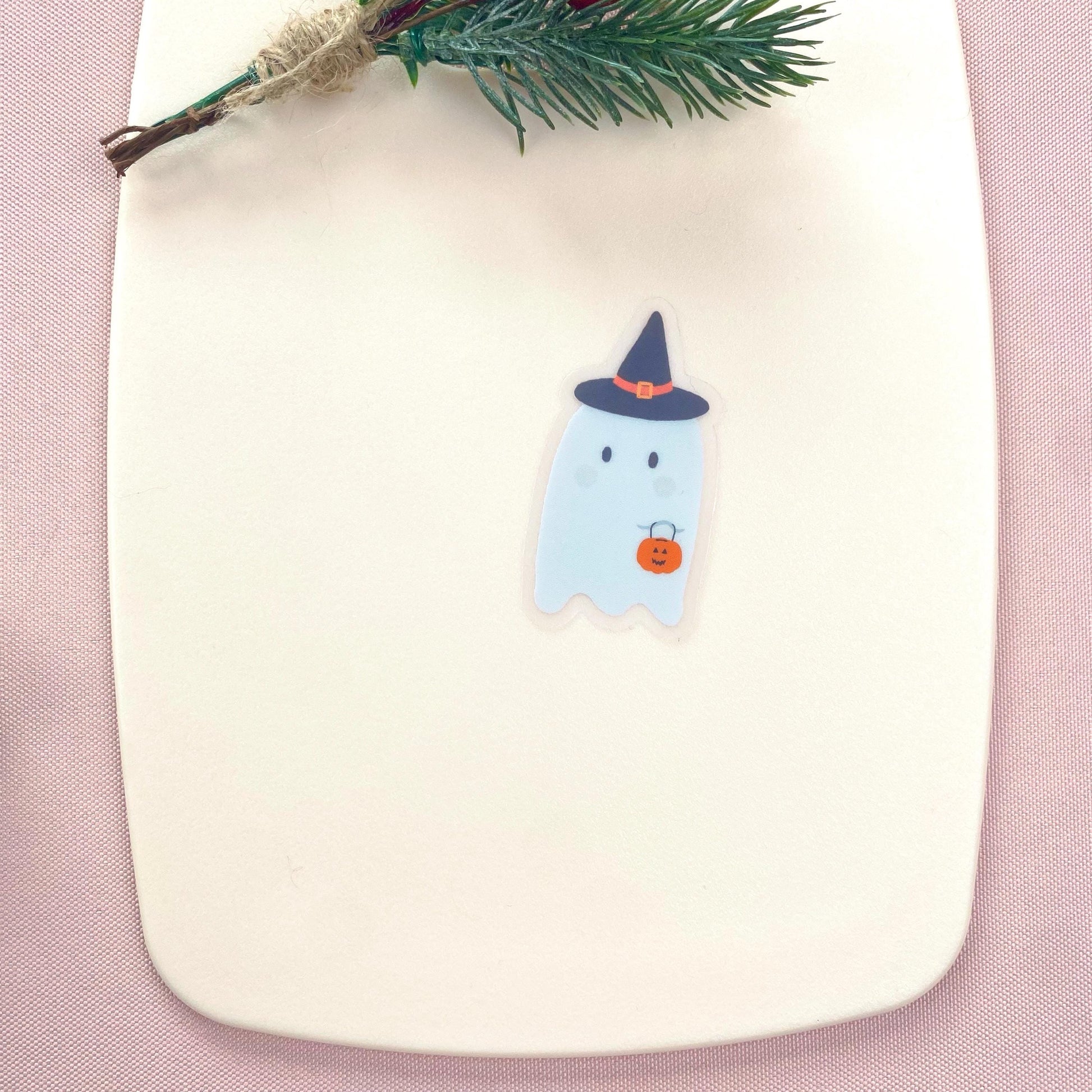 A sticker with a cartoon ghost wearing a witch hat and orange pumpkin on a pink background