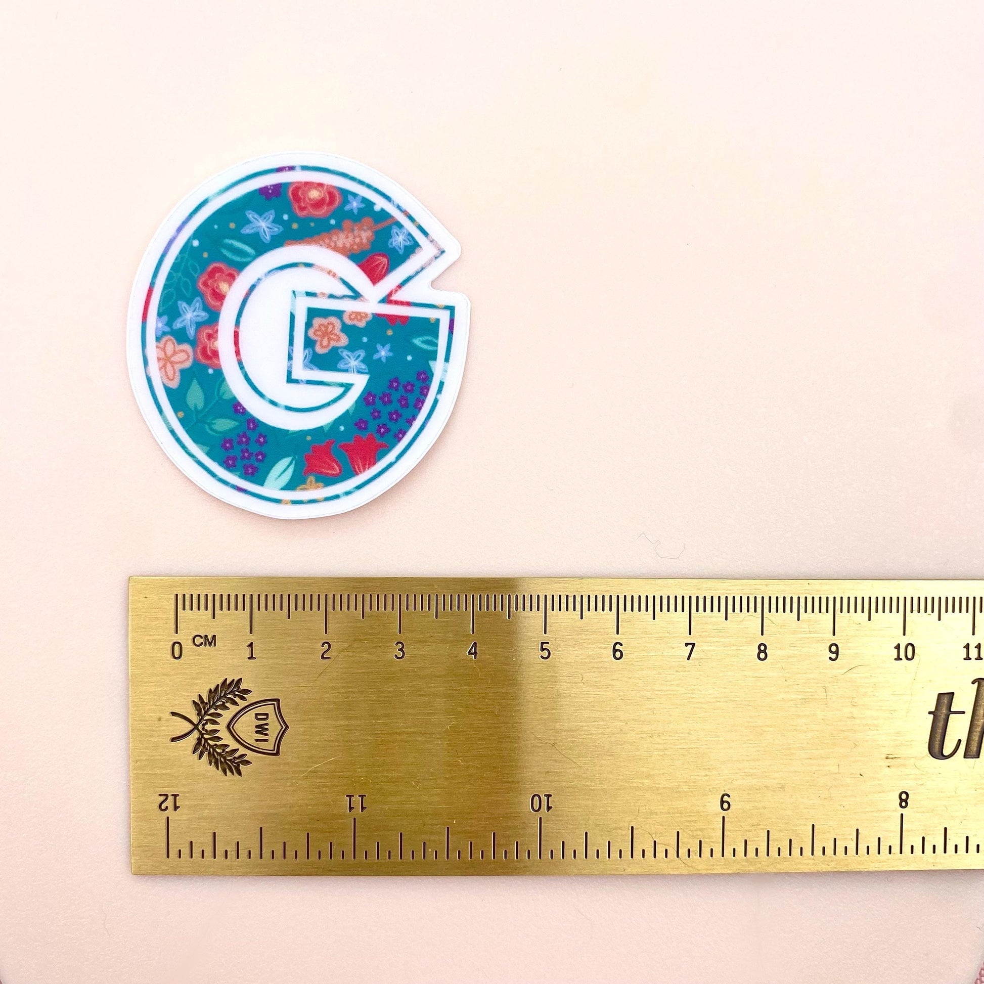 Decorative letter 'G' sticker with floral design next to a ruler on a beige background