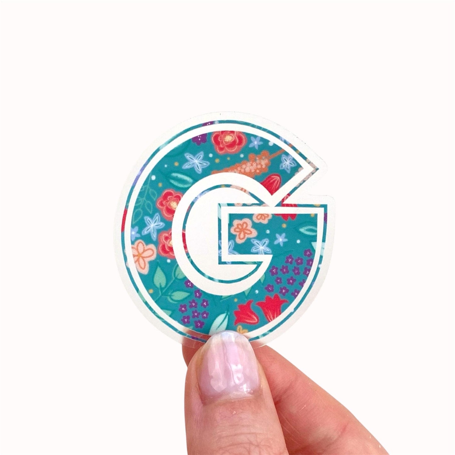 Hand holding a colourful letter 'G' sticker with floral design on a white background