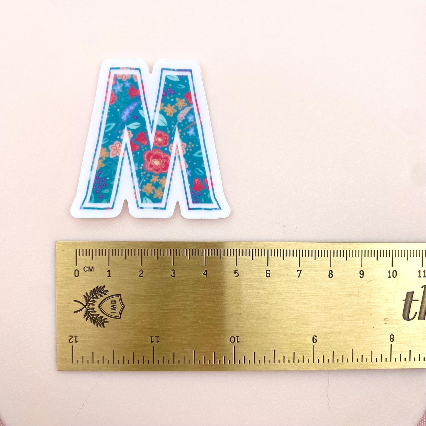 Floral letter 'M' sticker next to a ruler on a light pink background