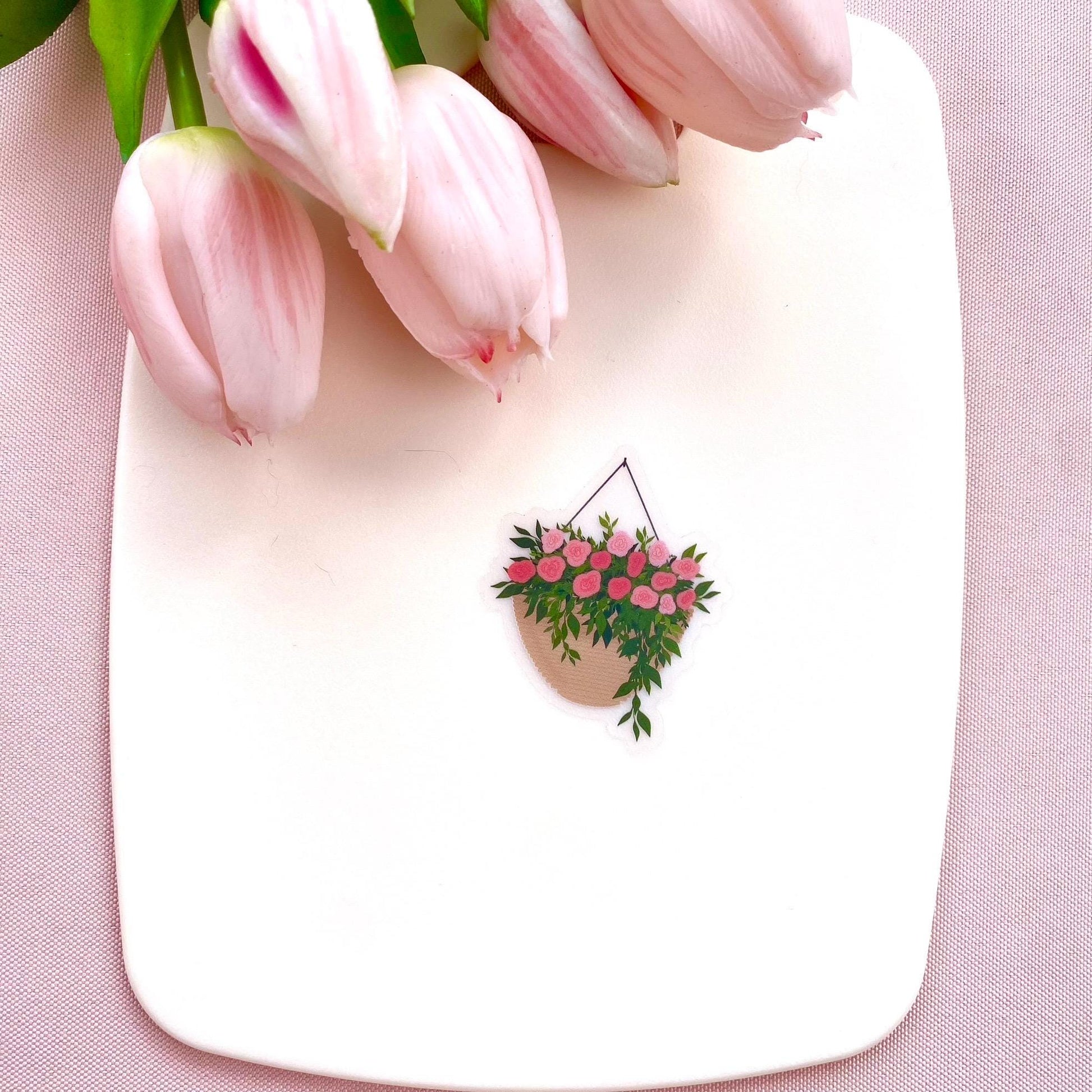 A sticker with a hanging planter with pink flowers against a white surface, with pink tulips in the background.