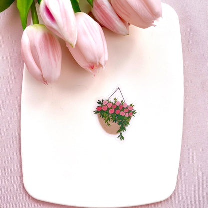 A sticker with a hanging planter with pink flowers against a white surface, with pink tulips in the background.