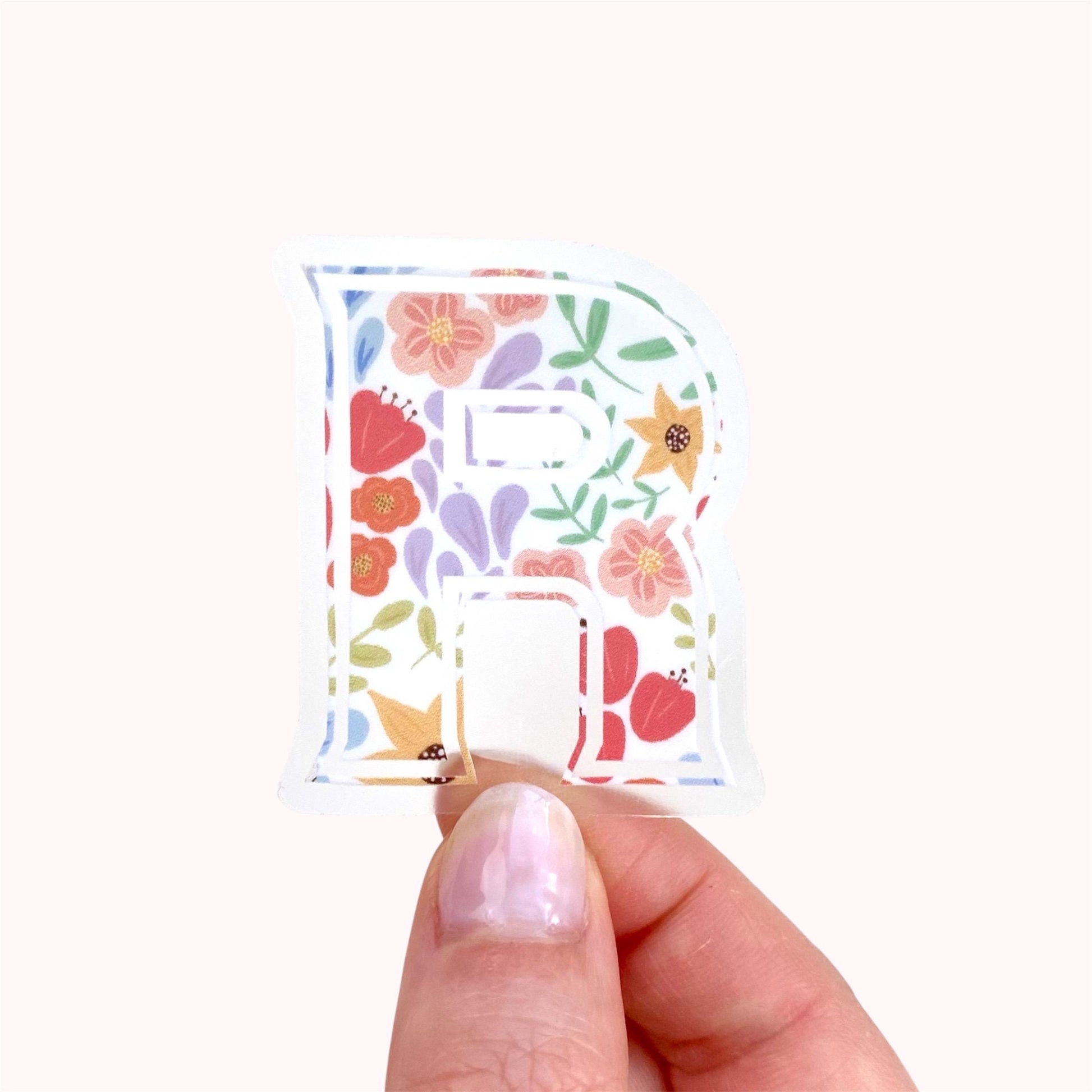 Hand holding a letter 'R' sticker with floral design on a white background