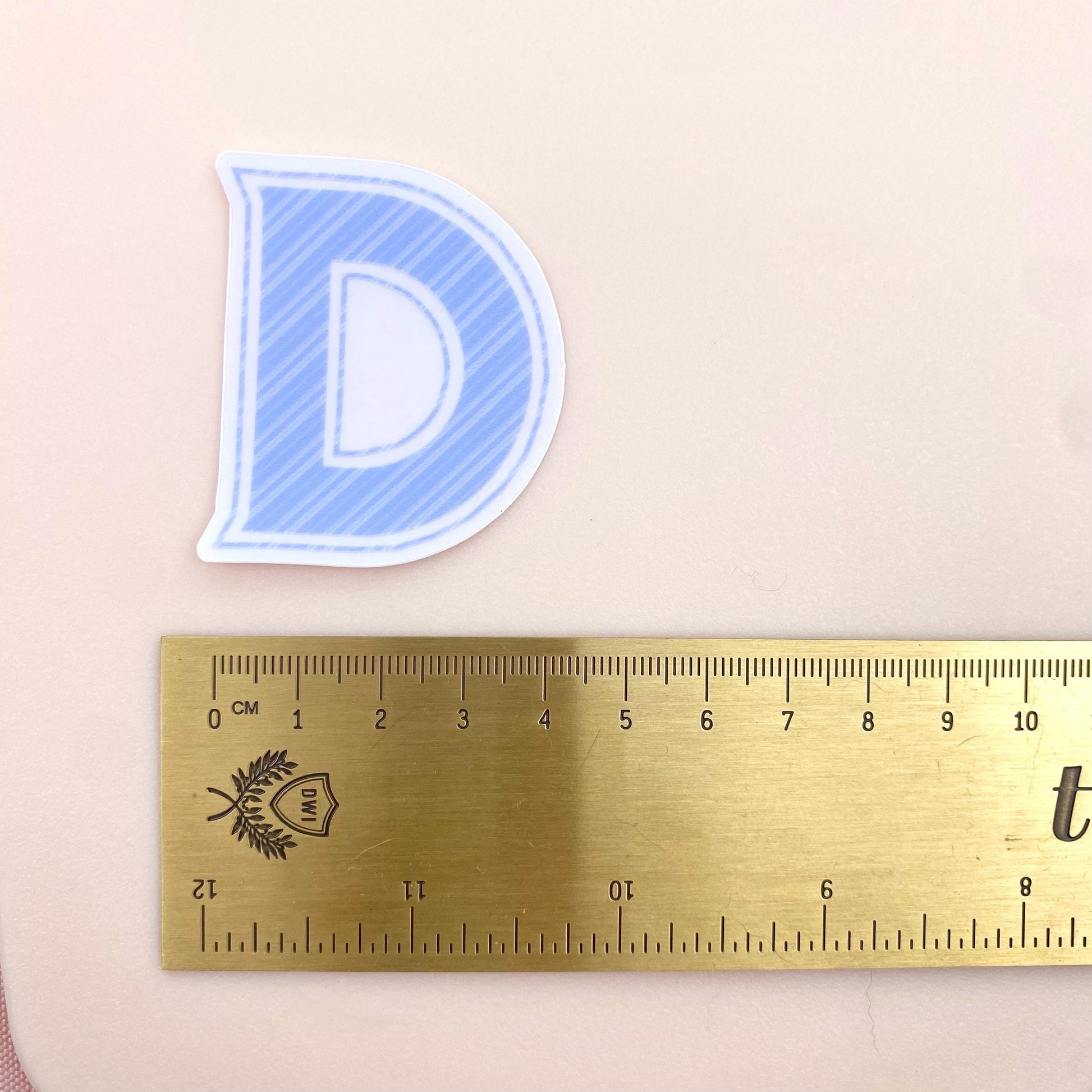 Blue striped letter 'D' sticker next to a ruler on a beige background