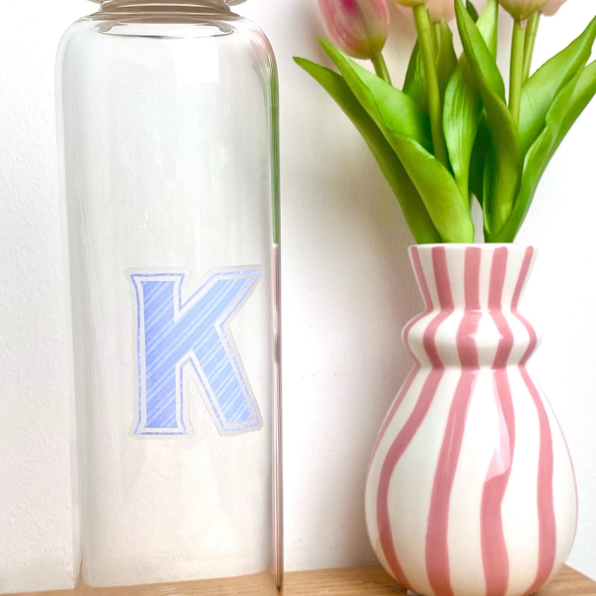 Clear bottle with blue letter 'K' next to a pink and white striped vase with tulips.