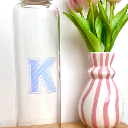 Clear bottle with blue letter 'K' next to a pink and white striped vase with tulips.