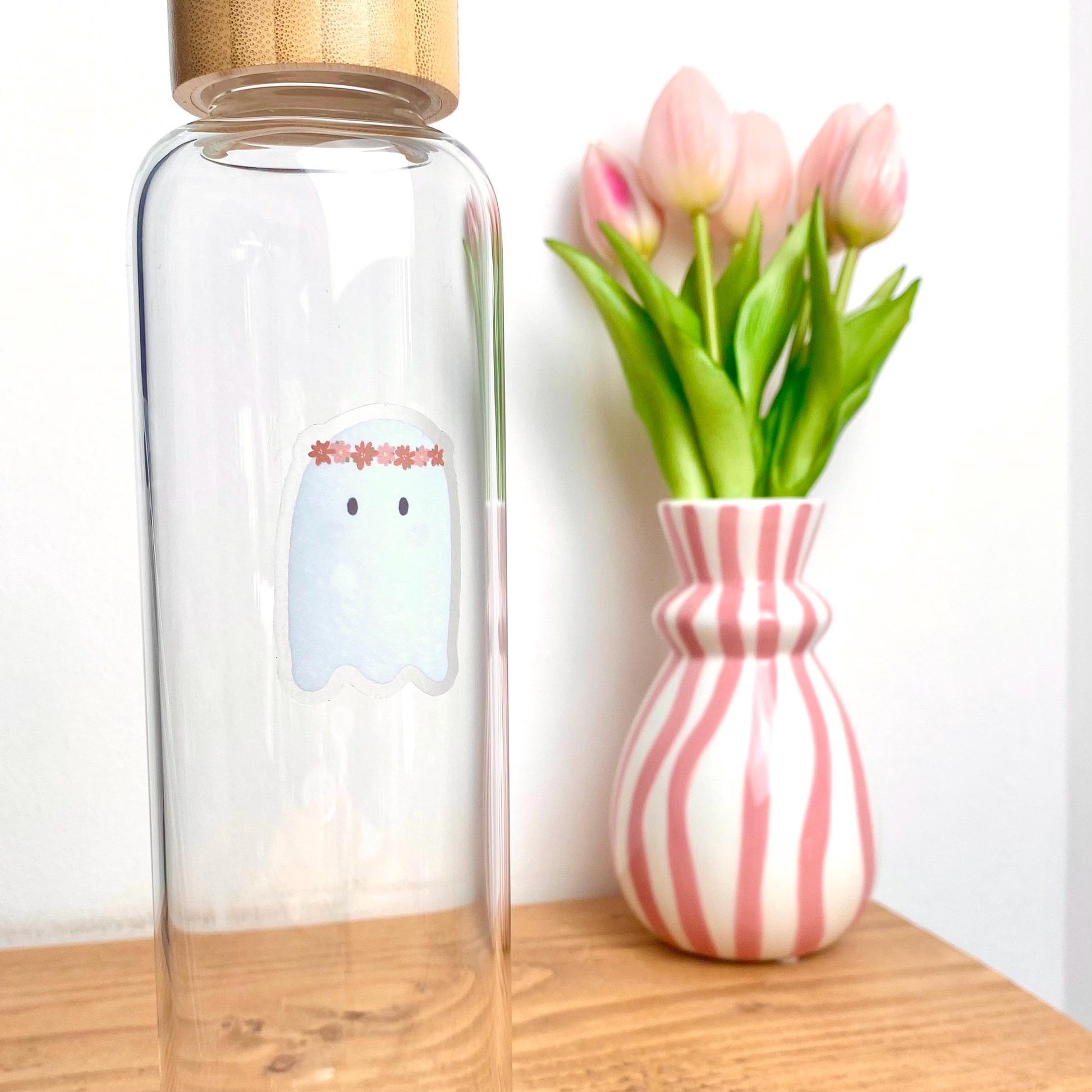 Clear glass bottle with a cartoon ghost sticker next to a pink and white striped vase with tulips on a wooden surface.
