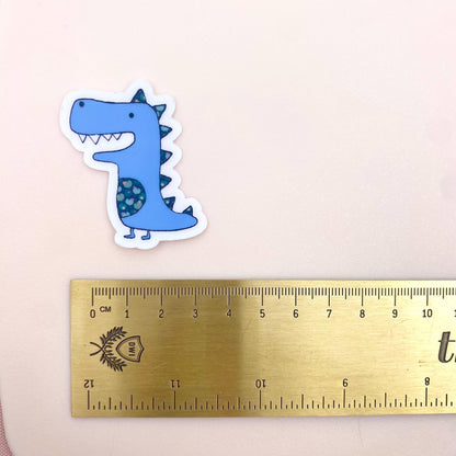 Blue dinosaur sticker next to a ruler on a pink background