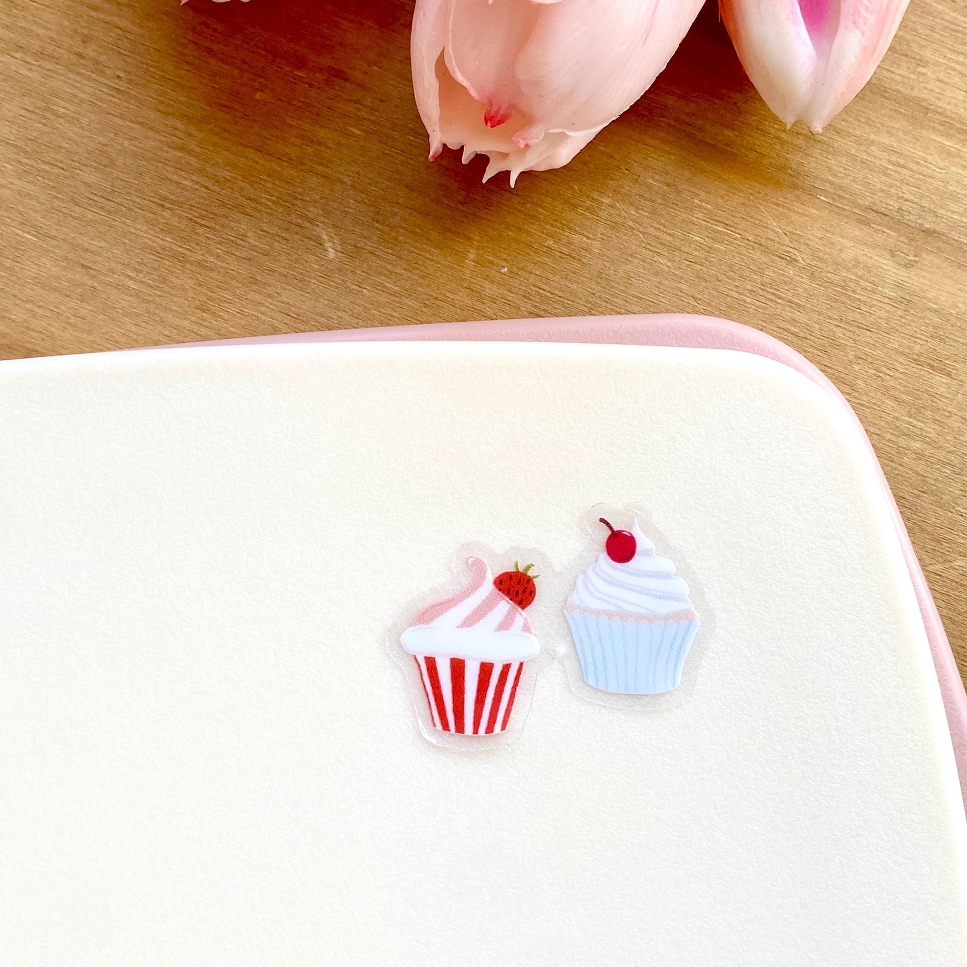 A sticker with two tiny cupcakes in pink and blue against a cream background.