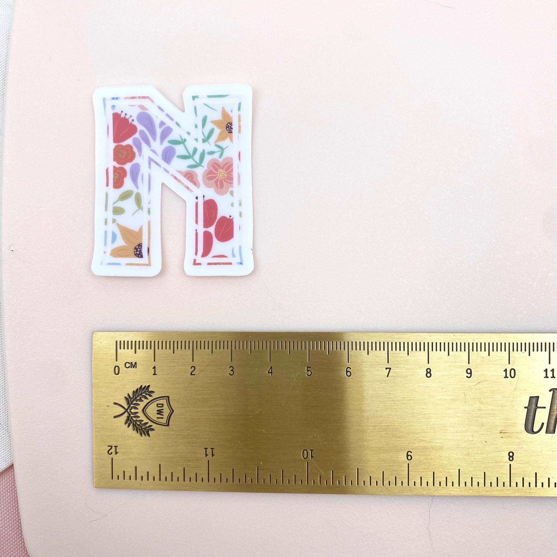 Decorative letter 'N' sticker with floral design next to a gold ruler on a pink background