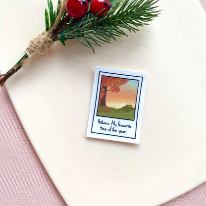 Sticker with polaroid design with autumn illustration and text on a white surface with greenery and berries.