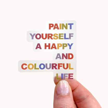 Hand holding a clear sticker with motivational quote 'Paint yourself a happy and colourful life' on a white background
