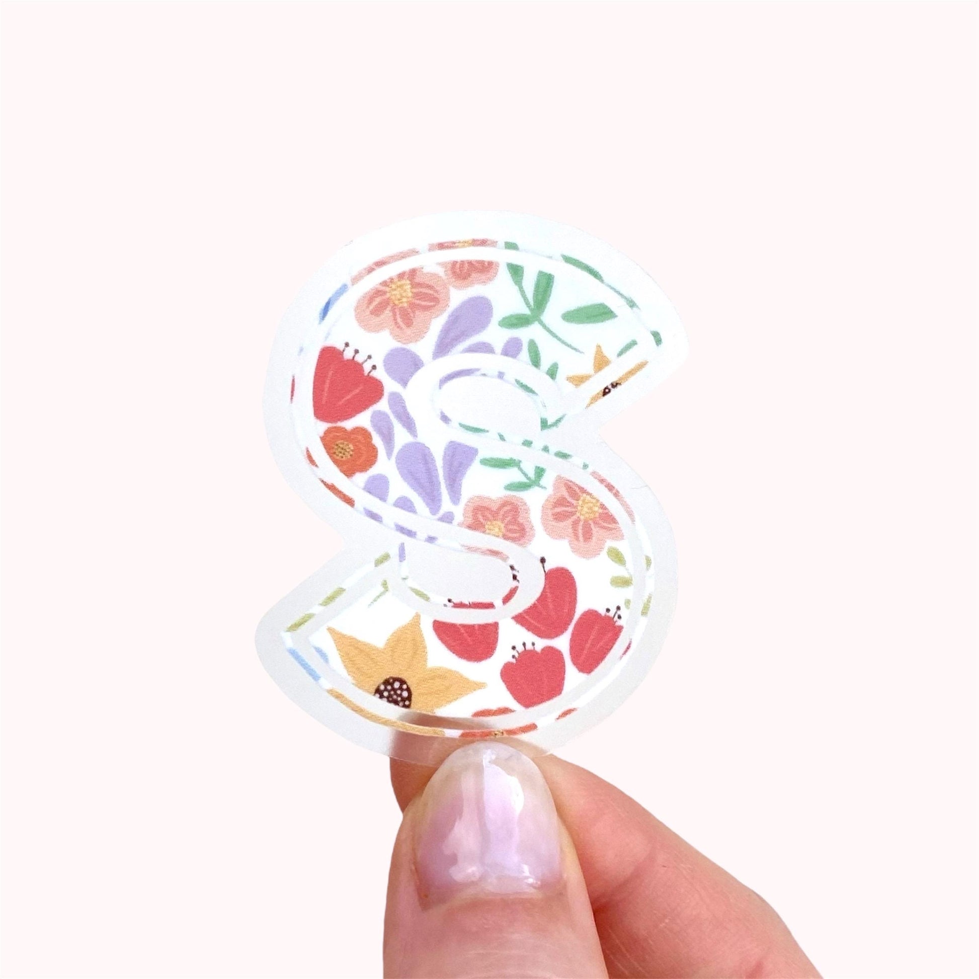 Decorative letter 'S' sticker with floral pattern held by hand against a pink background