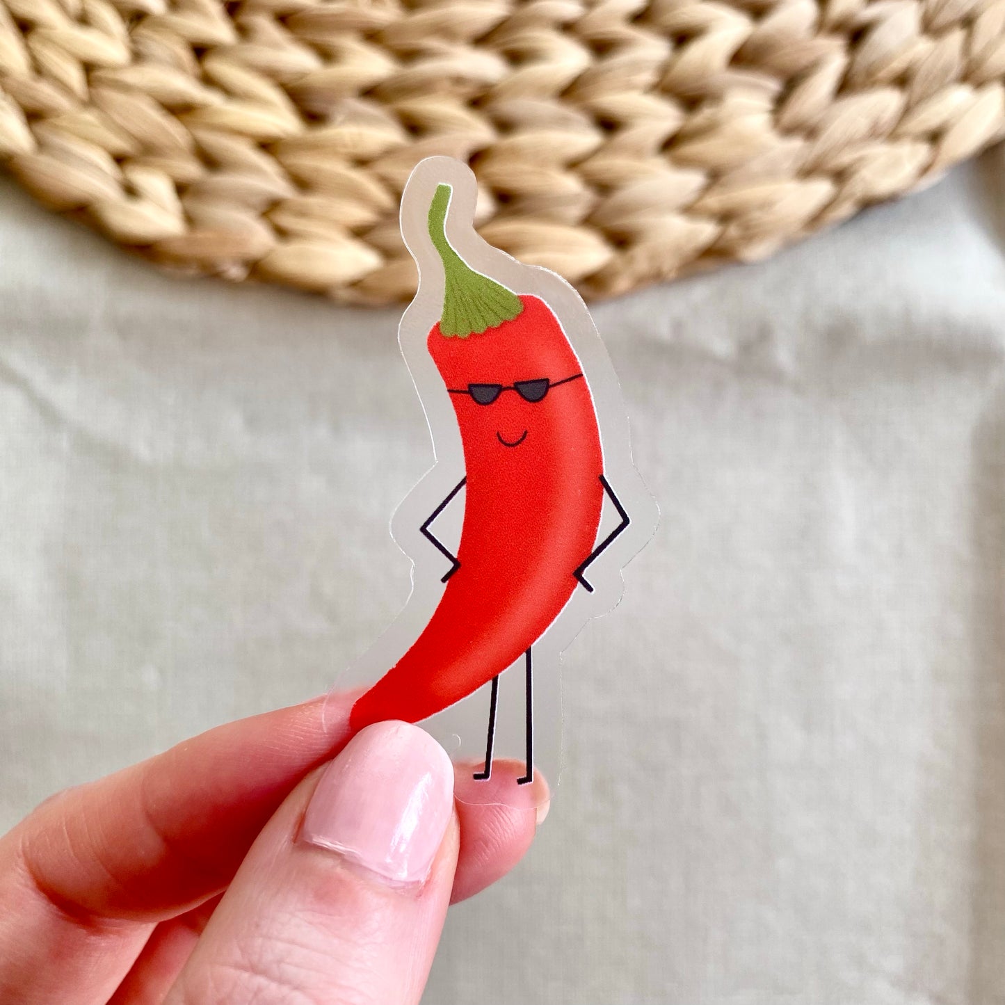 Hand holding a sticker of a red chili pepper with sunglasses against a neutral background