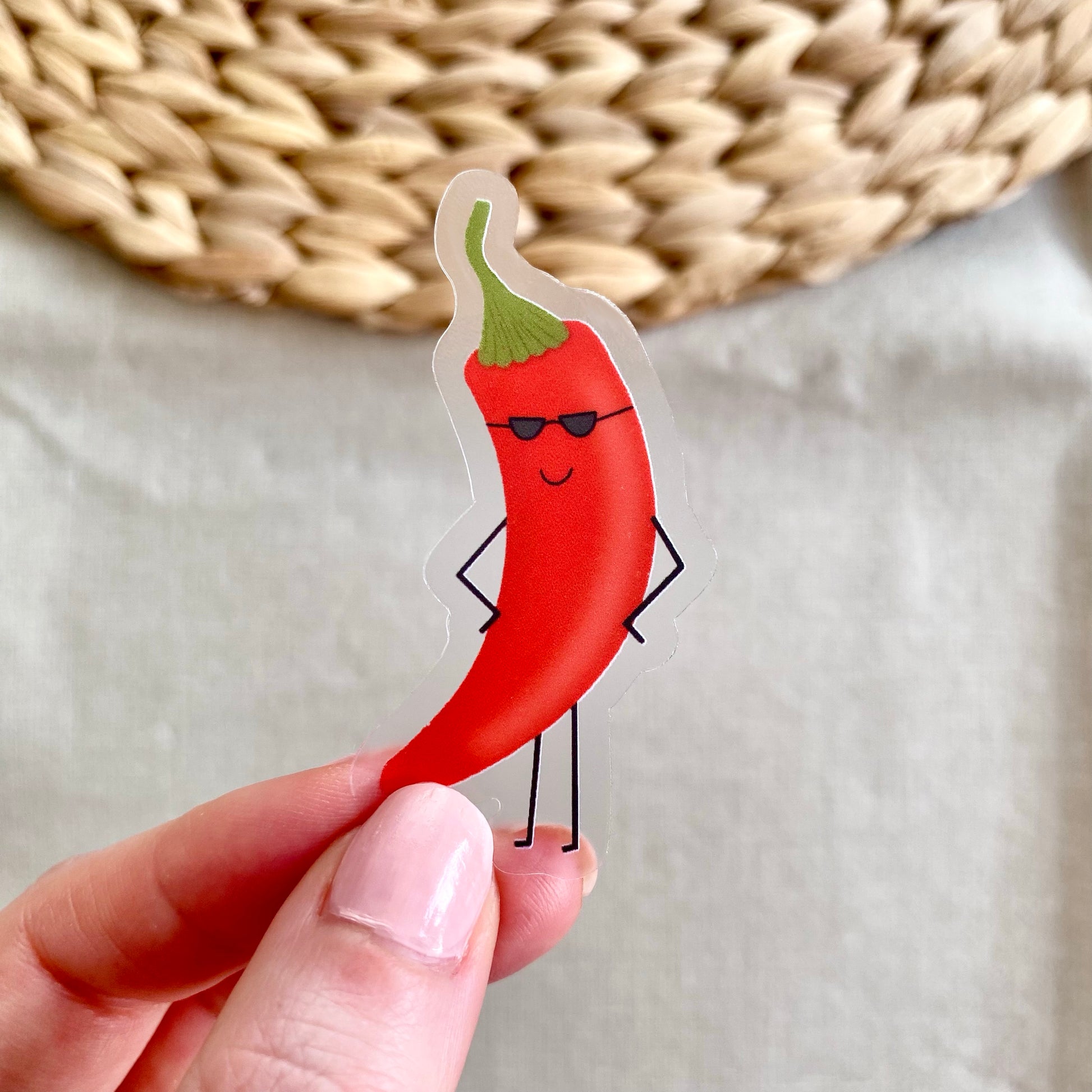 Hand holding a sticker of a red chili pepper with sunglasses against a neutral background