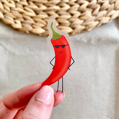 Hand holding a sticker of a red chili pepper with sunglasses against a neutral background