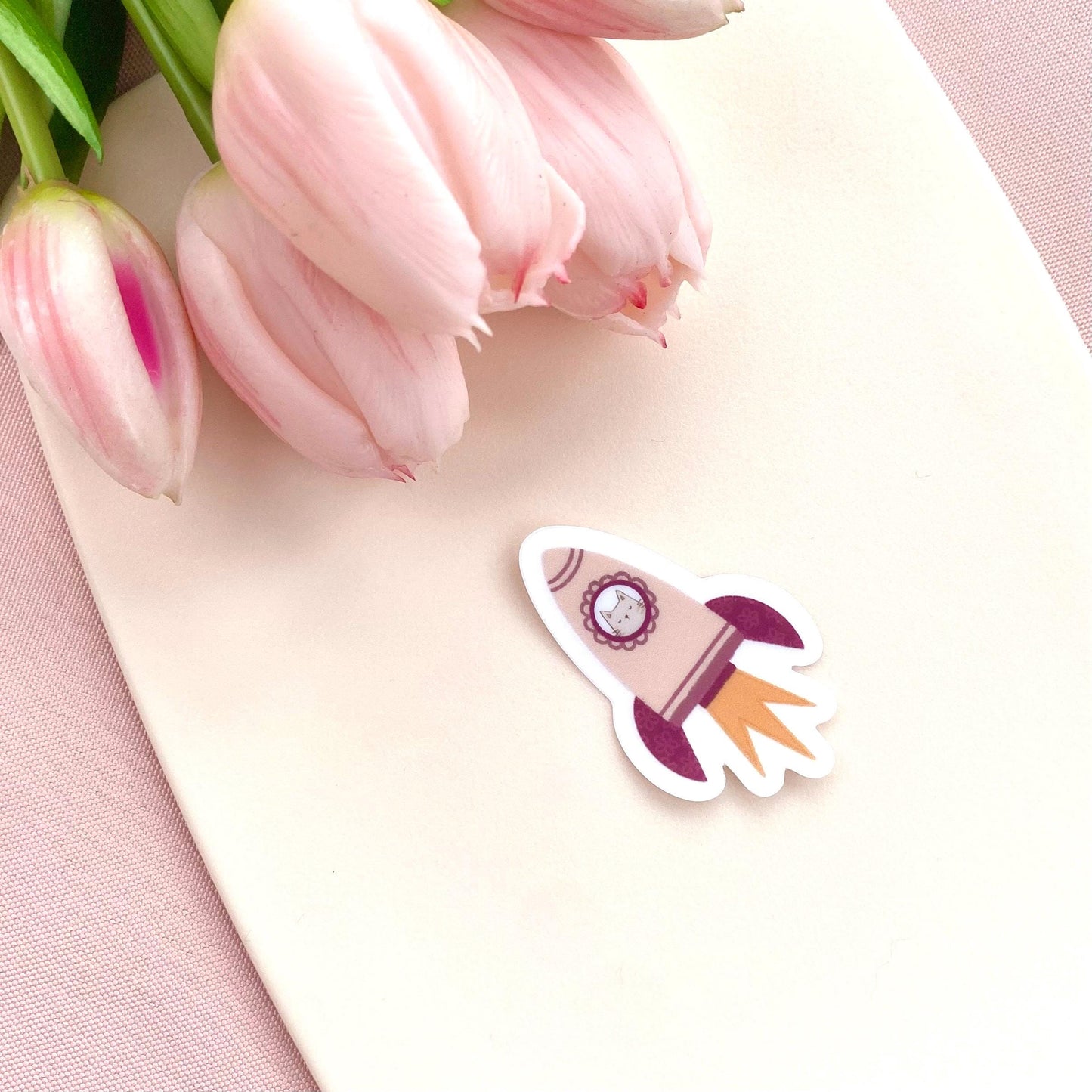 A pink rocket sticker with a grey cat in the window against a cream background with pink tulips