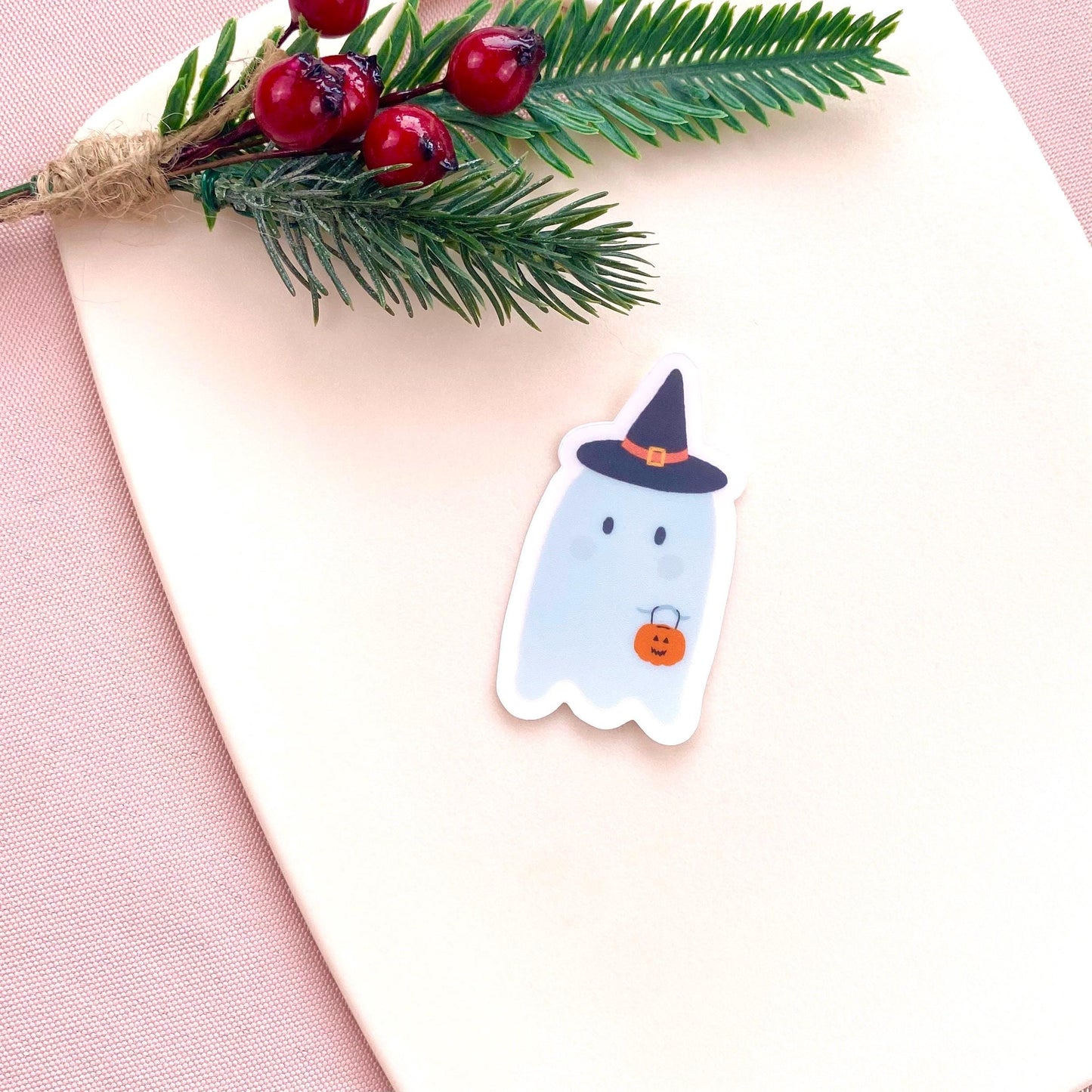 Sticker of a ghost with a witch hat on a pink surface with greenery