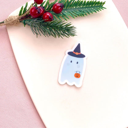 Sticker of a ghost with a witch hat on a pink surface with greenery