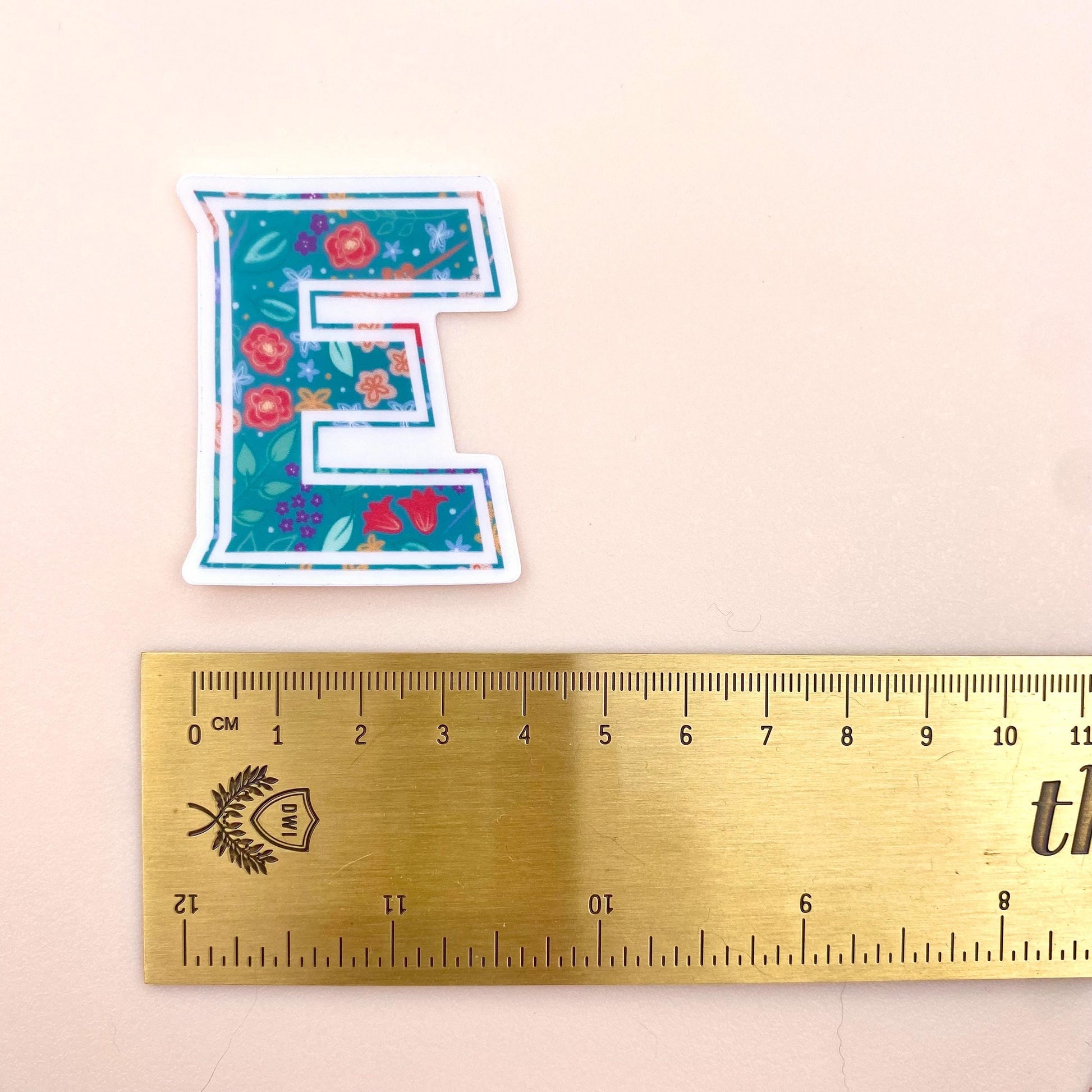 Floral letter 'E' sticker next to a ruler on a beige background