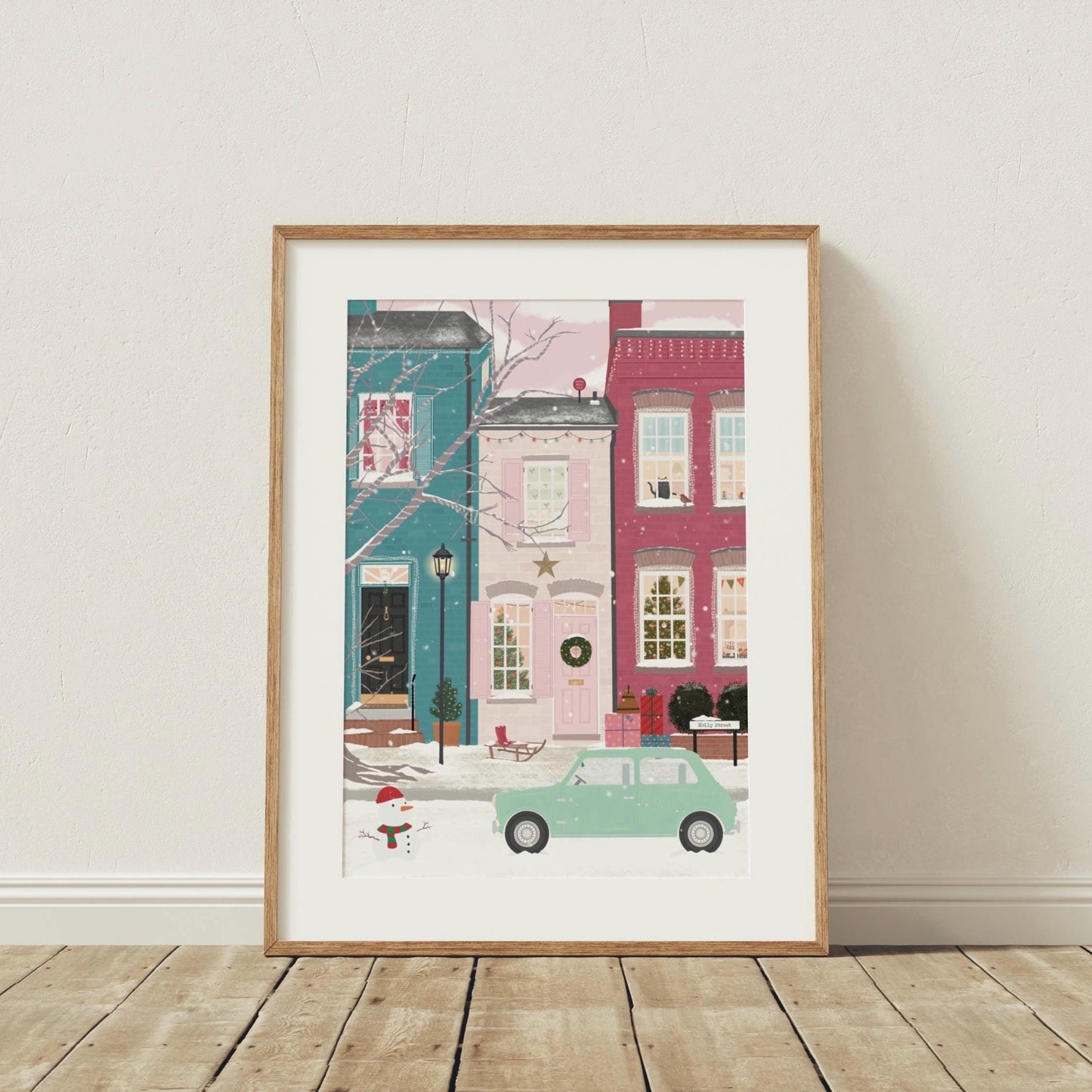 Framed artwork of a snowy street scene with houses and a car on a wooden floor.