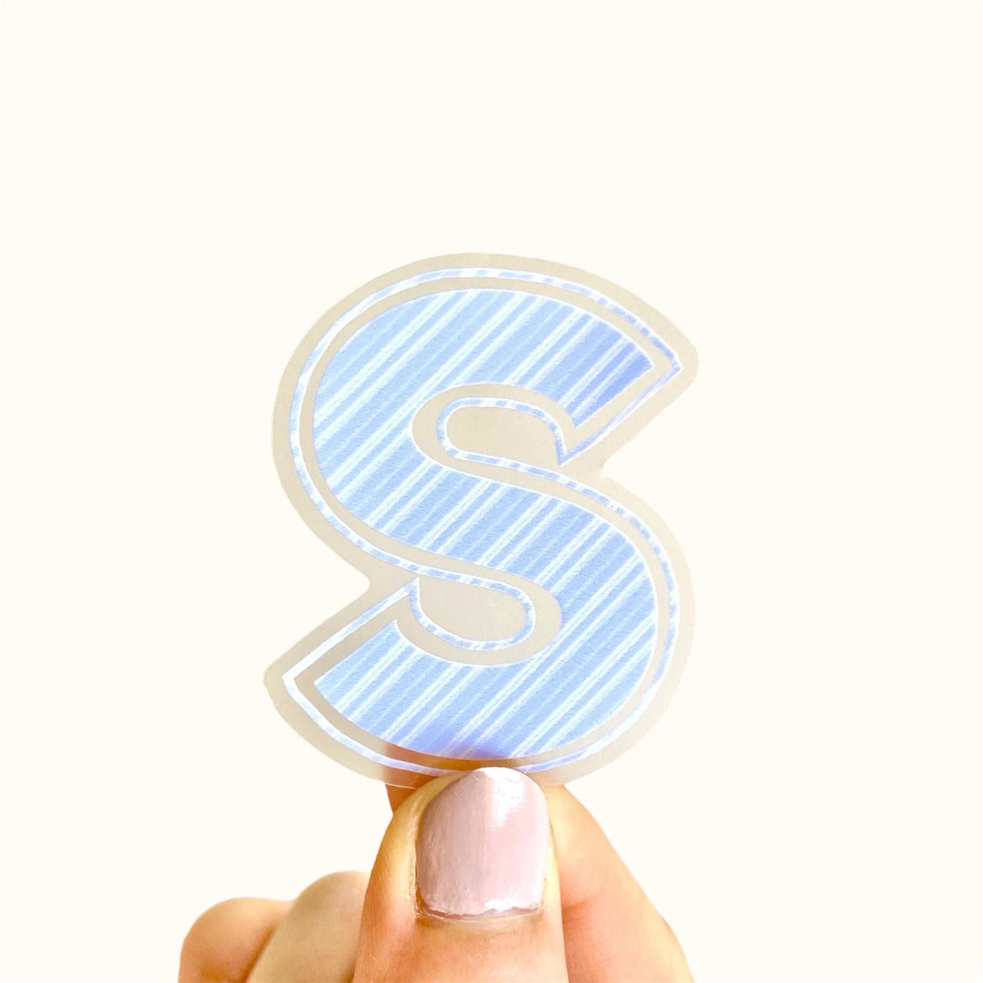 Hand holding a blue striped letter 'S' sticker against a white background