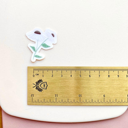 A clear sticker with daisies with white petals and green leaves on a white background with a gold ruler for scale.