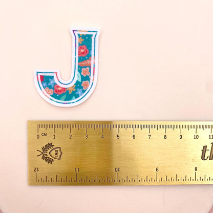 Floral letter 'J' sticker next to a ruler on a beige background