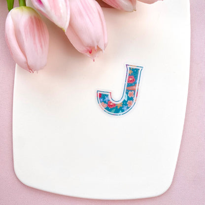Colourful letter 'J' sticker on a pink background with tulips.