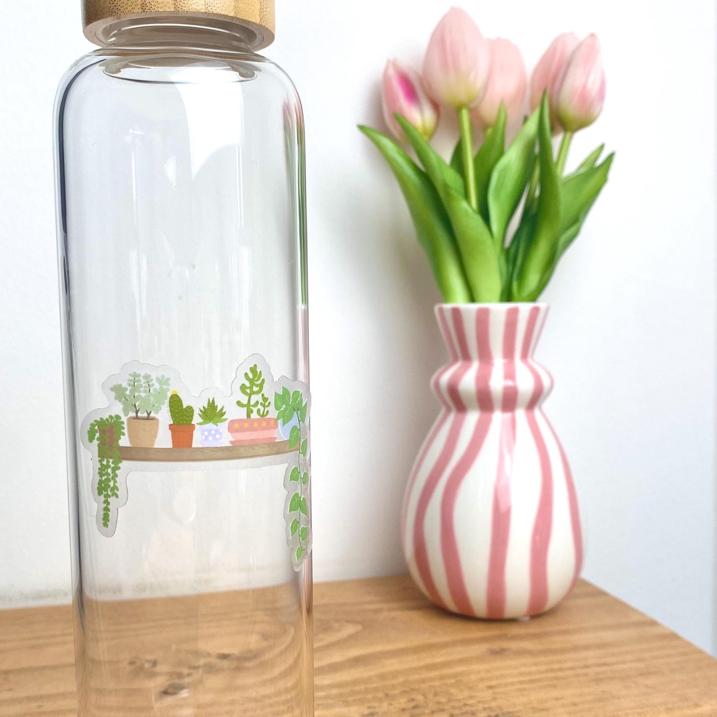Clear glass bottle with plant-themed sticker next to a striped vase with tulips on a wooden surface.