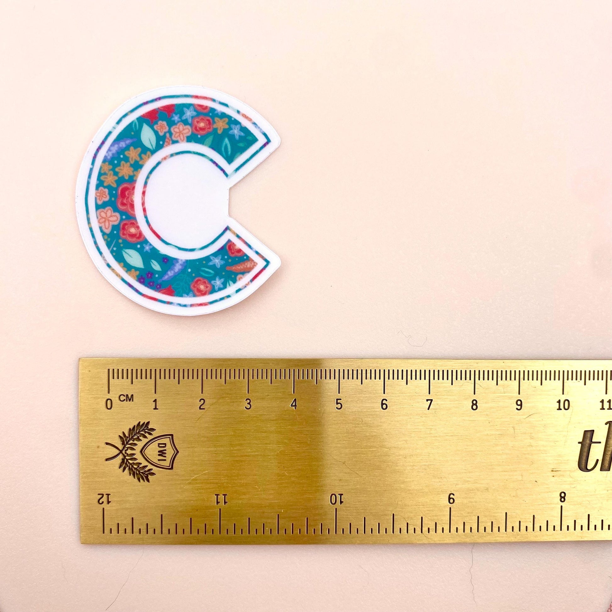 Floral letter 'C' sticker next to a ruler on a beige background