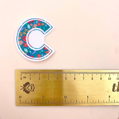 Floral letter 'C' sticker next to a ruler on a beige background