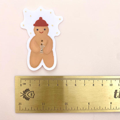 Sticker of a gingerbread man holding a festive hot chocolate mug next to a ruler for scale on a beige background