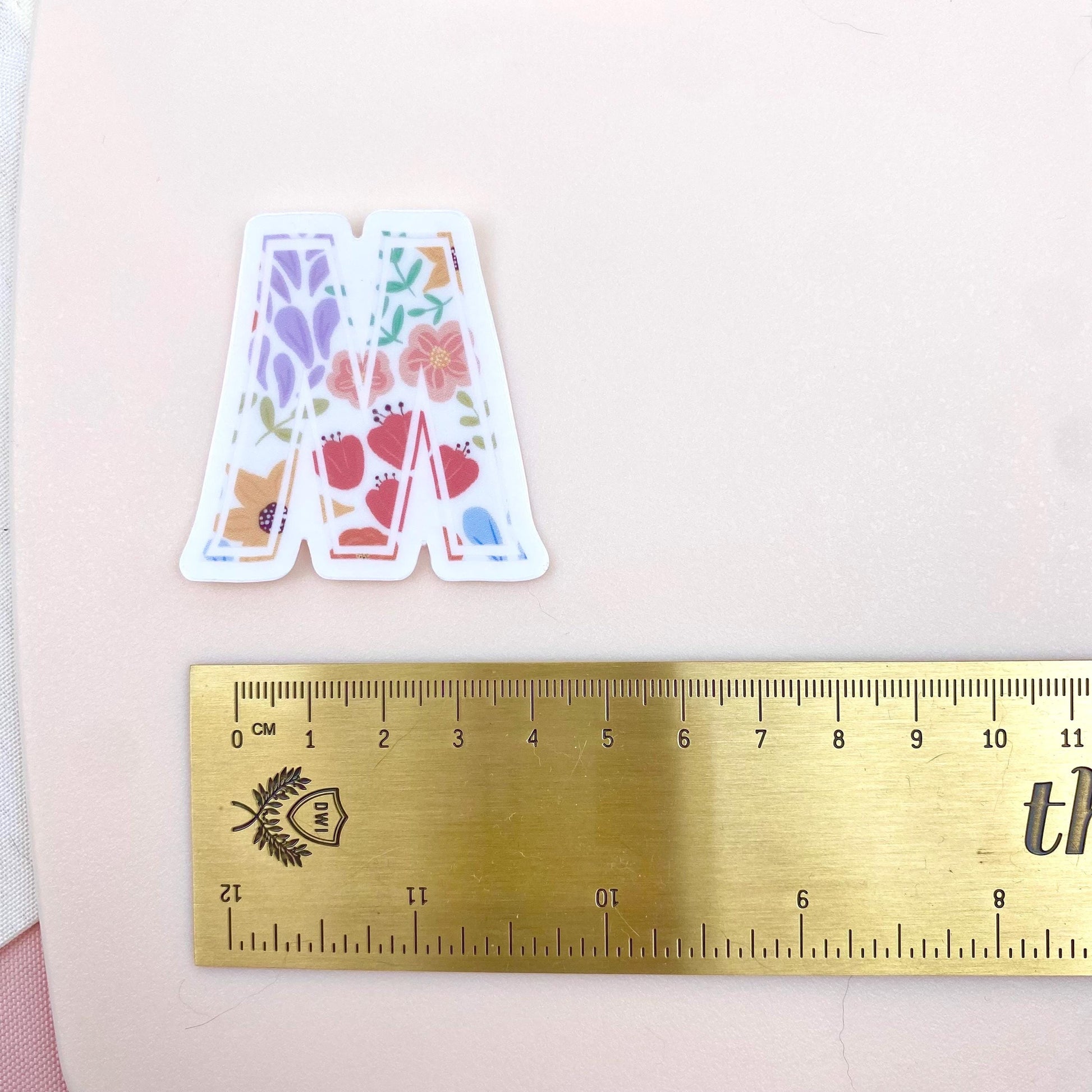 Decorative letter 'M' sticker with floral patterns on a white background with a gold ruler