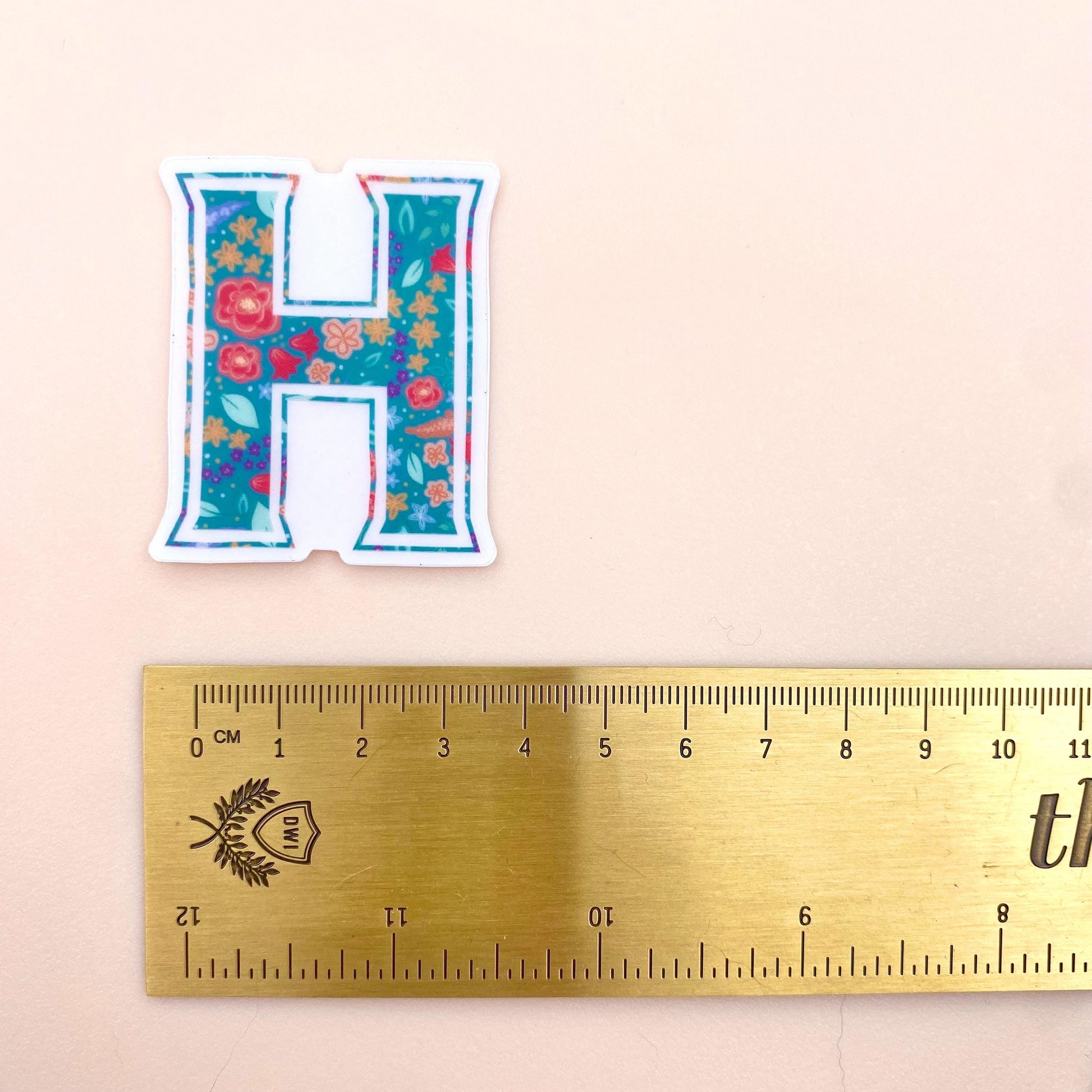 Floral letter 'H' sticker next to a ruler on a beige background