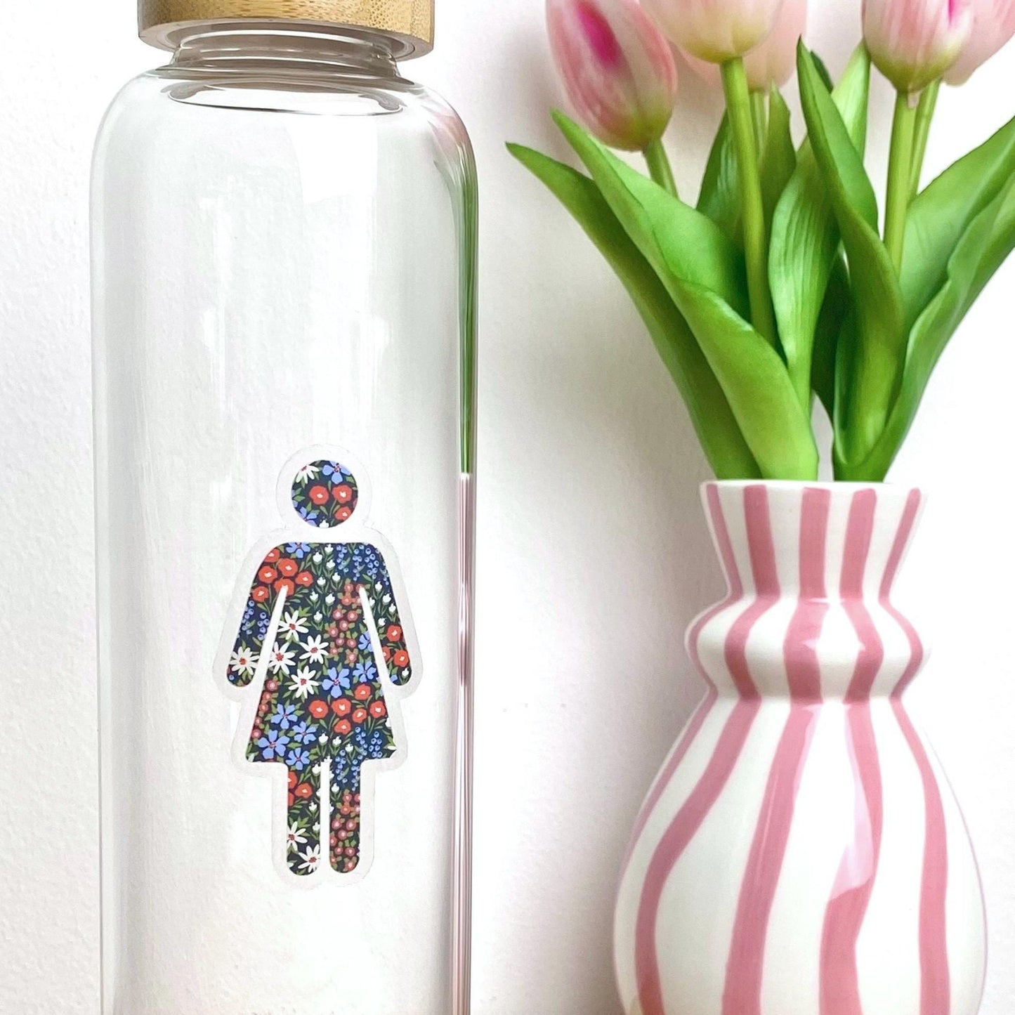 Clear glass bottle with floral woman symbol design next to a striped vase with tulips on a white background
