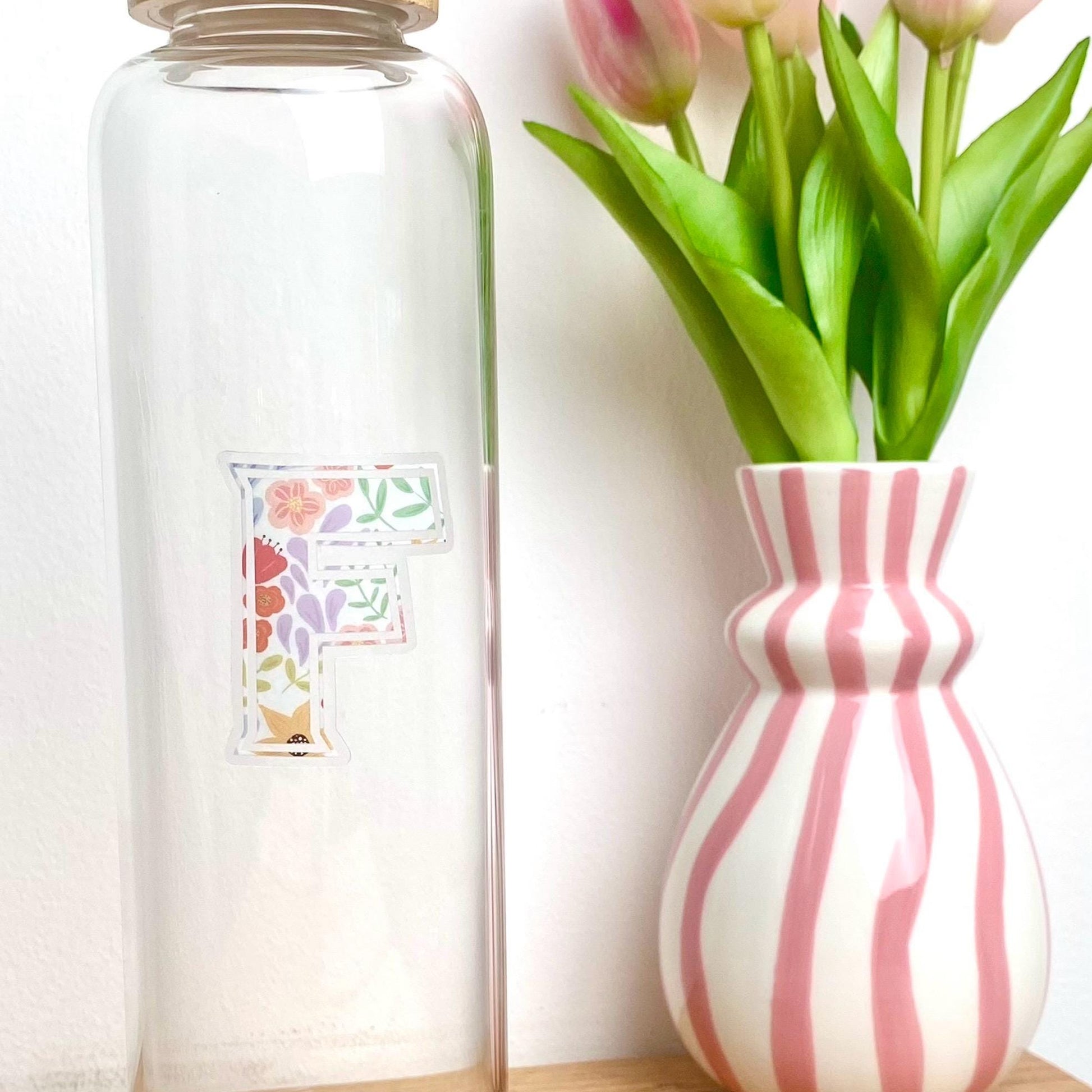 Clear glass bottle with a floral letter 'F' clear sticker next to a pink and white striped vase with tulips.