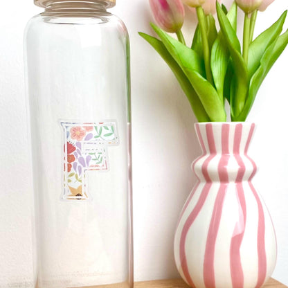 Clear glass bottle with a floral letter 'F' clear sticker next to a pink and white striped vase with tulips.