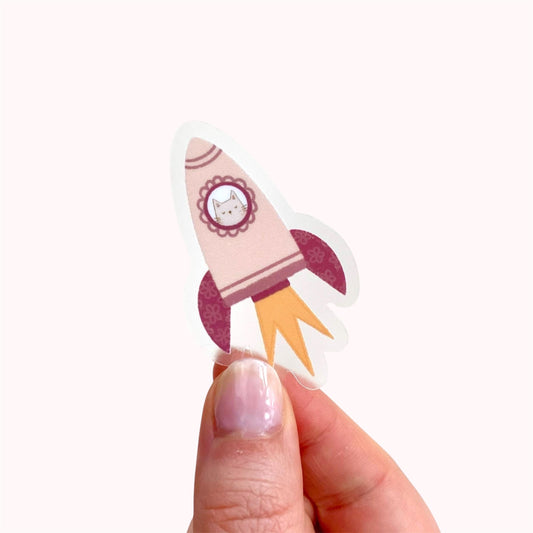 Hand holding a sticker of a pink rocket with a grey cat in the window