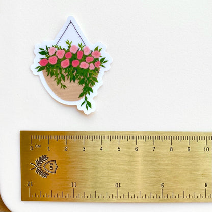 A sticker with a hanging planter with pink flowers against a white background with a gold ruler for scale.