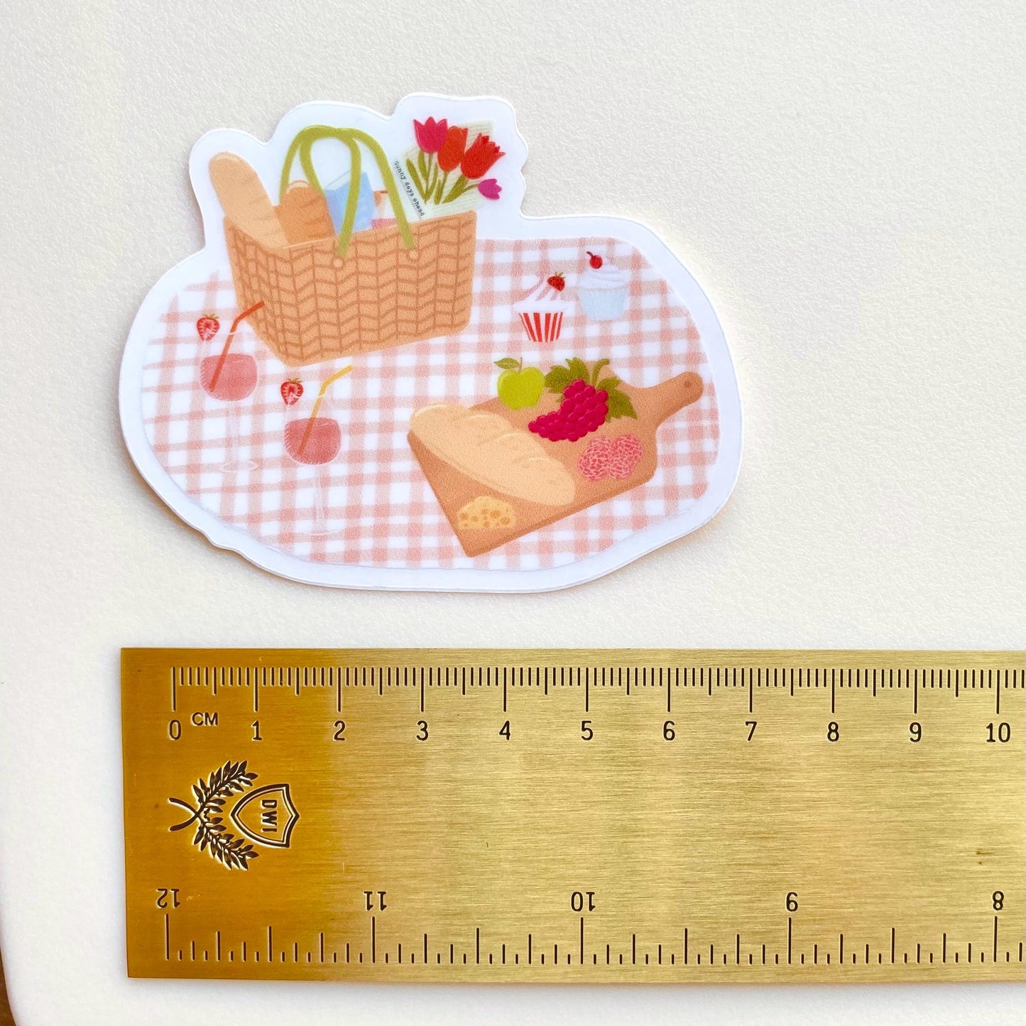 Sticker with picnic scene on a checkered blanket next to a ruler for scale