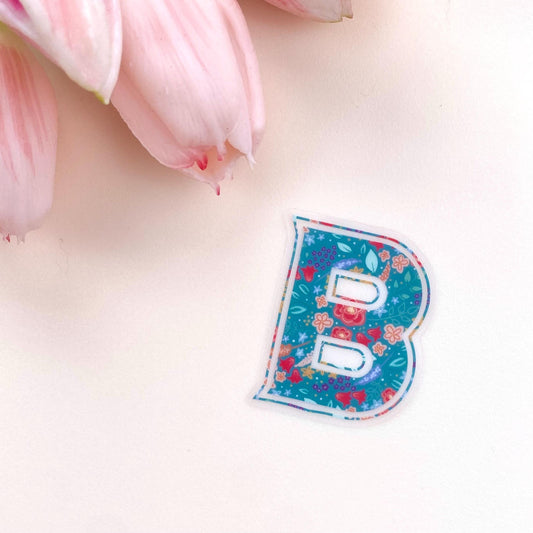 Clear sticker with a teal floral decorated letter 'B' against a cream background.